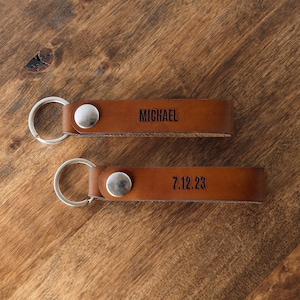 May include: Two brown leather keychains with silver key rings. The top keychain has the name "MICHAEL" engraved on it. The bottom keychain has the date "7.12.23" engraved on it.