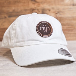 May include: White baseball cap with a curved brim and adjustable strap. A circular brown leather patch on the front displays the initials "ajp" in a stylized font. The cap is made of a soft fabric.