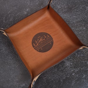 May include: A square, brown leather catchall tray with a circular emblem in the center. The emblem features a mountain and tree design with the text "NW CUSTOM MADE". The tray has snap closures at each corner.