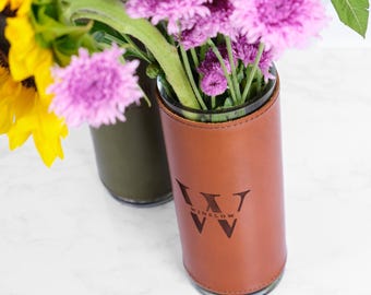 May include: A brown leather vase with the word "Winslow" and a logo etched on the front. The vase holds purple and white flowers. A second vase with green leather is visible in the background. The flowers include sunflowers.