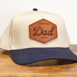 May include: A beige and navy blue baseball cap with a brown leather patch that says "Dad Established MMXXV".
