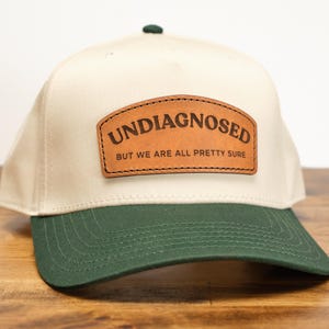 May include: A beige baseball cap with a green brim. The front of the cap features a brown leather patch with the text "UNDIAGNOSED BUT WE ARE ALL PRETTY SURE".