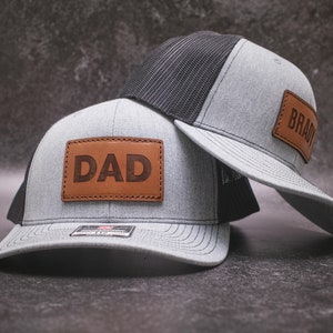 Może przedstawiać: Dwie szare i czarne czapki trucker z brązowymi skórzanymi naszywkami. Naszywka na przedniej czapce ma napis "DAD", a naszywka na tylnej czapce ma napis "BRADY".