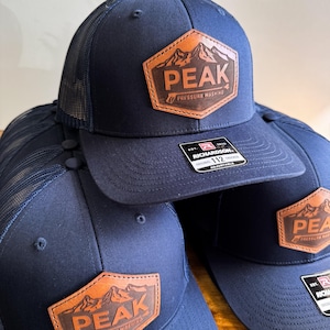 Puede incluir: Tres gorras de camionero azul marino con parches de cuero marrón que representan una cordillera y la palabra "PEAK" con el texto "Pressure Washing" debajo. Las gorras están apiladas una encima de la otra. La gorra de abajo tiene una etiqueta blanca con el texto "Richardson 112" en ella.