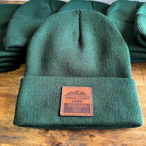 Peut inclure: Un bonnet en tricot vert foncé avec un patch en cuir marron qui dit "Your Logo Here Established 2025".