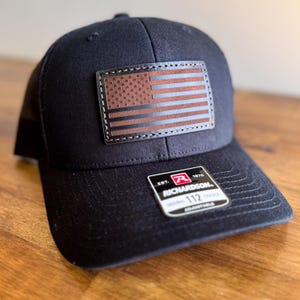 Puede incluir: Gorra de camionero negra con un parche de cuero marrón con la bandera estadounidense en la parte delantera. La gorra tiene una parte trasera de malla negra y una correa ajustable negra con una etiqueta "Richardson 112".