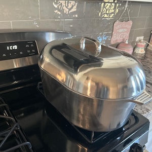 May include: A large, shiny stainless steel roasting pan with a lid sits on a black gas stove. The pan has a handle on the lid and a rack on the side. The stove is in a kitchen setting.