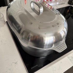 May include: A shiny, stainless steel oval roasting pan with a matching lid and handle. The pan sits on a black Samsung stovetop. The lid has a rectangular handle. The pan is designed for cooking and baking.