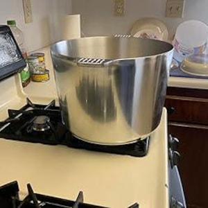 May include: A large, shiny, stainless steel pot with two handles sits on a black stovetop burner. The pot is cylindrical with a wide opening. Other kitchen items, including a paper towel roll and dishes, are visible in the background.