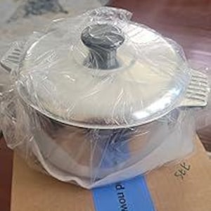 May include: A new, stainless steel cooking pot with a lid, packaged in clear plastic. The pot has two side handles and a dark gray knob on the lid. It sits on a cardboard box with a blue tape strip.