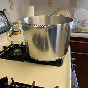 May include: A large, shiny, stainless steel pot with two handles sits on a black stovetop burner. The pot is cylindrical with a wide opening. Other kitchen items, including a paper towel roll and dishes, are visible in the background.