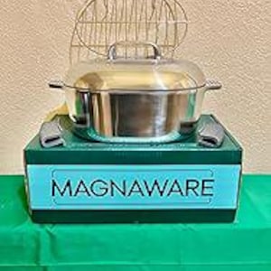 May include: A stainless steel MagnaWare cooking pot with a lid and handles, sitting on a green and black box. A metal rack is behind the pot. The box has the word "MAGNAWARE" in white letters.