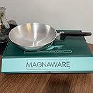May include: A stainless steel frying pan with a black handle rests on a teal-colored appliance. The pan is round and has a silver interior. The appliance has the word "MAGNAWARE" printed on the front.