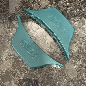 May include: Two teal-colored, curved silicone pot handle covers with the word MAGNAWARE embossed in a rectangular frame on the front surface. These kitchen accessories feature a smooth, contoured design intended for gripping hot cookware handles.