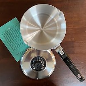 May include: A stainless steel saucepan with a long black handle and a matching base. The pan has a shiny, reflective interior and is sitting on a dark wooden surface. A green paper with text is to the left of the pan.