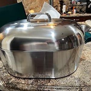 May include: A stainless steel oval roaster with a lid and a handle. The roaster has green handles on each side. The roaster is sitting on a speckled countertop.