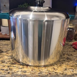May include: A large, shiny stainless steel pot with a lid and teal handles. The pot has a black knob on the lid and reflects the surrounding environment. This cookware is suitable for cooking various dishes.