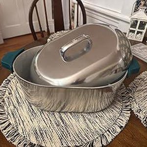 May include: A stainless steel oval casserole dish with a matching lid and teal handles. The dish sits on a round, cream-colored placemat with a black fringe. A wooden chair and other decorative items are in the background.