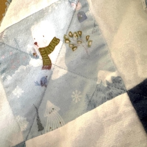 May include: A patchwork quilt featuring a winter scene with a snowman wearing a green scarf, snowflakes, and trees. The quilt has a blue and white color scheme, with a ruffled edge.