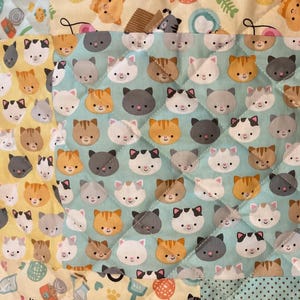 May include: A patchwork quilt featuring a repeating pattern of cat faces in various colors, including orange, gray, white, and brown. The quilt has a light blue background with a diamond pattern, and other sections with cat-themed prints.