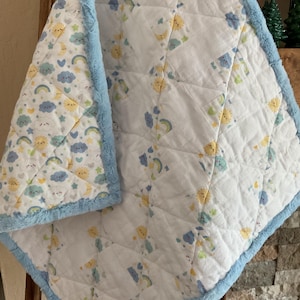 May include: A white baby blanket with a pattern of yellow suns, blue clouds, and rainbows. The blanket has a light blue plush border. The blanket is folded over a wooden surface.