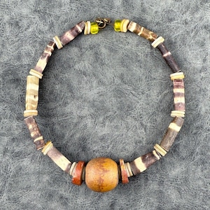 May include: A handcrafted necklace featuring a large, round, light brown bead as a focal point. The necklace is composed of various beads in shades of brown, cream, and purple, with small yellow beads near the clasp. The necklace is designed for a natural, earthy aesthetic.