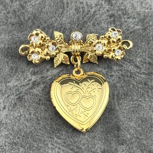 May include: Gold-toned brooch featuring a floral design with clear rhinestone accents. The brooch has a heart-shaped locket pendant with an engraved design of two hearts. A classic and elegant piece of jewelry.