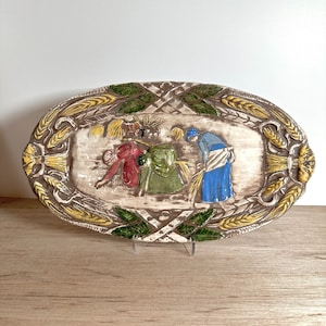 May include: An oval ceramic serving dish with a raised design of three figures harvesting wheat. The dish is decorated with wheat stalks, leaves, and a crosshatch pattern in brown, yellow, green, and blue.