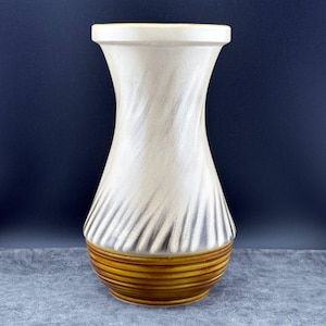 May include: A ceramic vase with a cream-colored upper section featuring a textured, swirling design. The base is a warm brown with horizontal stripes. The vase has a wide top and a narrower base.