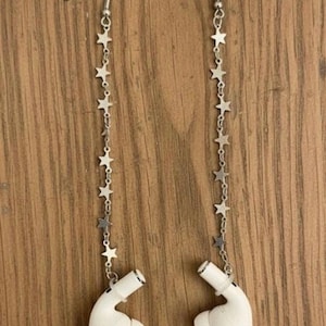 Peut inclure: Une paire d'écouteurs sans fil blancs avec des chaînes d'étoiles argentées. Les écouteurs sont suspendus à des crochets d'oreilles argentés. Les boucles d'oreilles sont présentées sur un fond en bois.