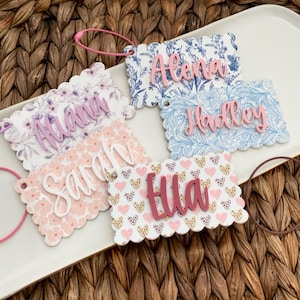 May include: A collection of personalized name tags with scalloped edges. Each tag features a different floral or heart pattern and a name in a contrasting color. The tags are attached to pink loops and are arranged on a white tray.