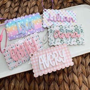 May include: A collection of personalized name tags with scalloped edges and various patterns. The tags feature names like "Isla," "Lilian," "Anna," "Emily," and "Avery" in cursive font. Each tag has a different background, including pastel gradients, floral prints, and gingham.