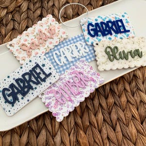 May include: A collection of personalized name tags with scalloped edges. Each tag features a different name in a contrasting color, such as "Gabriel" in navy blue or "Olivia" in green. The tags have various patterned backgrounds, including floral and star designs.