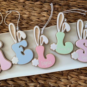 May include: Five wooden bunny-shaped ornaments with different colored letters and names. The letters are A, E, L, S, and the names are "William Sky", "Layla", and "Sarah".