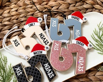 May include: A collection of personalized Christmas ornaments shaped like the number "67". Each ornament features a name, a Santa hat, and the year "'25". The ornaments are in various colors and patterns, including glitter and checkered designs.