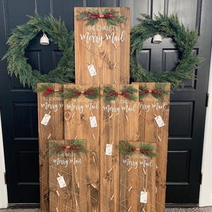 May include: Six wooden signs with a rustic finish, each with a different name and the words "Merry Mail" written in white cursive. The signs are decorated with sprigs of greenery and red berries. The signs are hung on a wall with a black door in the background.