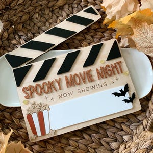 May include: A wooden film clapperboard sign featuring the text SPOOKY MOVIE NIGHT in glittery orange letters. The board includes a white rectangular space for writing, a popcorn bucket graphic, two black bat cutouts, and small star accents.
