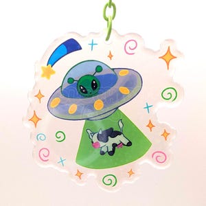 Peut inclure: Un porte-clés en acrylique transparent avec une illustration de dessin animé représentant un extraterrestre vert dans un OVNI gris et bleu. L'OVNI émet un rayon tracteur vert soulevant une petite vache noire et blanche avec des étoiles colorées.