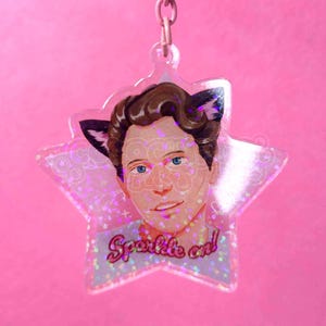 May include: A pink star-shaped keychain with a portrait of a person with cat ears. The portrait has blue eyes and brown hair. The keychain has the words "Sparkle on!" in pink script.