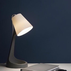 May include: A modern gray desk lamp with a white, pleated lampshade is angled towards a gray notebook and a black computer mouse. The lamp has a curved base and a slender neck. The background is a dark blue.