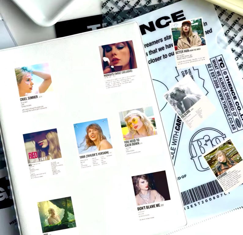 May include: A white sheet of paper with several colorful square stickers featuring album covers by Taylor Swift. The stickers feature album titles like "Cruel Summer", "Red", "You Need to Calm Down", "1989 (Taylor's Version)", "Don't Blame Me", "Folklore", "Evermore", and "Better Man".