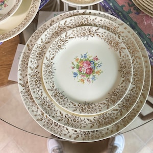 Puede incluir: Tres platos de cerámica blanca con motivos florales dorados. Los platos están anidados, con el plato más pequeño en el centro que presenta un ramo de flores rosas, amarillas y azules.