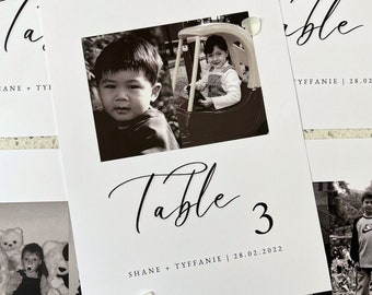 May include: A white rectangular table number card featuring a black and white photograph of two children, one sitting in a toy car. The text reads Table 3, SHANE + TYFFANIE | 28.02.2022 in a black script and sans-serif font, decorated with small white flower petals.