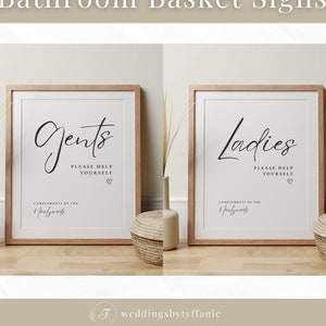 May include: Two framed signs with a light brown wood frame and white background. The signs have black calligraphy text that reads "Gents Please Help Yourself Complements of the Newlyweds" and "Ladies Please Help Yourself Complements of the Newlyweds".
