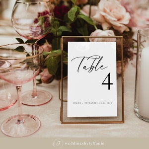 May include: A rectangular table number card held in a thin gold metal frame. The white card features the word Table in black script, the number 4 in a large black font, and the text SHANE + TYFFANIE | 28.02.2022 printed in a smaller black font.
