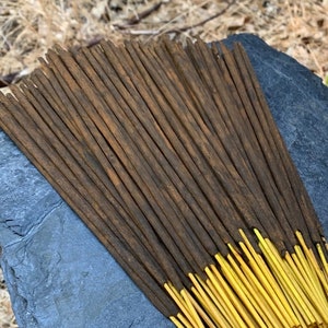 May include: A bundle of brown and yellow incense sticks arranged on a gray stone. The sticks are stacked on a tree stump in a natural outdoor setting.