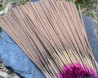 Può includere: Un fascio di bastoncini di incenso con steli marroni e punte rosa acceso, appoggiato su una pietra grigio scuro. I bastoncini di incenso sono disposti a ventaglio, con le punte rosa che creano un contrasto vibrante.