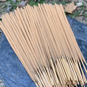 May include: A collection of light brown incense sticks, displayed on a dark grey stone. The incense sticks are neatly arranged and parallel. The tips of the incense sticks are a lighter shade of brown. The background shows fallen leaves.
