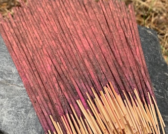 May include: A bundle of incense sticks with a reddish-brown color and a natural wood base. The sticks are arranged in a fan shape on a gray stone surface.