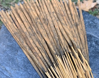 Peut inclure: Un ensemble de bâtonnets d'encens bruns clairs avec des extrémités foncées, posés sur une pierre grise foncée. Les bâtonnets d'encens sont disposés en éventail, pointant vers le haut. L'arrière-plan est flou, avec des feuilles mortes.